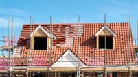 Scaffold poles surrounding the partly tiled roof with dormer windows of a new bungalow being built, against a blue sky.