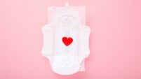 Bright red heart on opened white sanitary towel with wings on light pink table background. Pastel color. Closeup. Top down view.