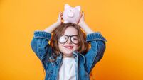 Cute little girl in eyeglasses shaking piggy box on orange background