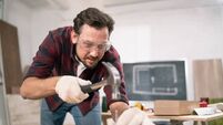 iStock-1308388224.jpg Hammering a nail into board. A carpenter wearing a red flannel shirt, jeans and cloth protective gloves nails the wooden boards