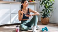 Athletic woman eating a healthy bowl of muesli with fruit sitting on floor in the kitchen at home